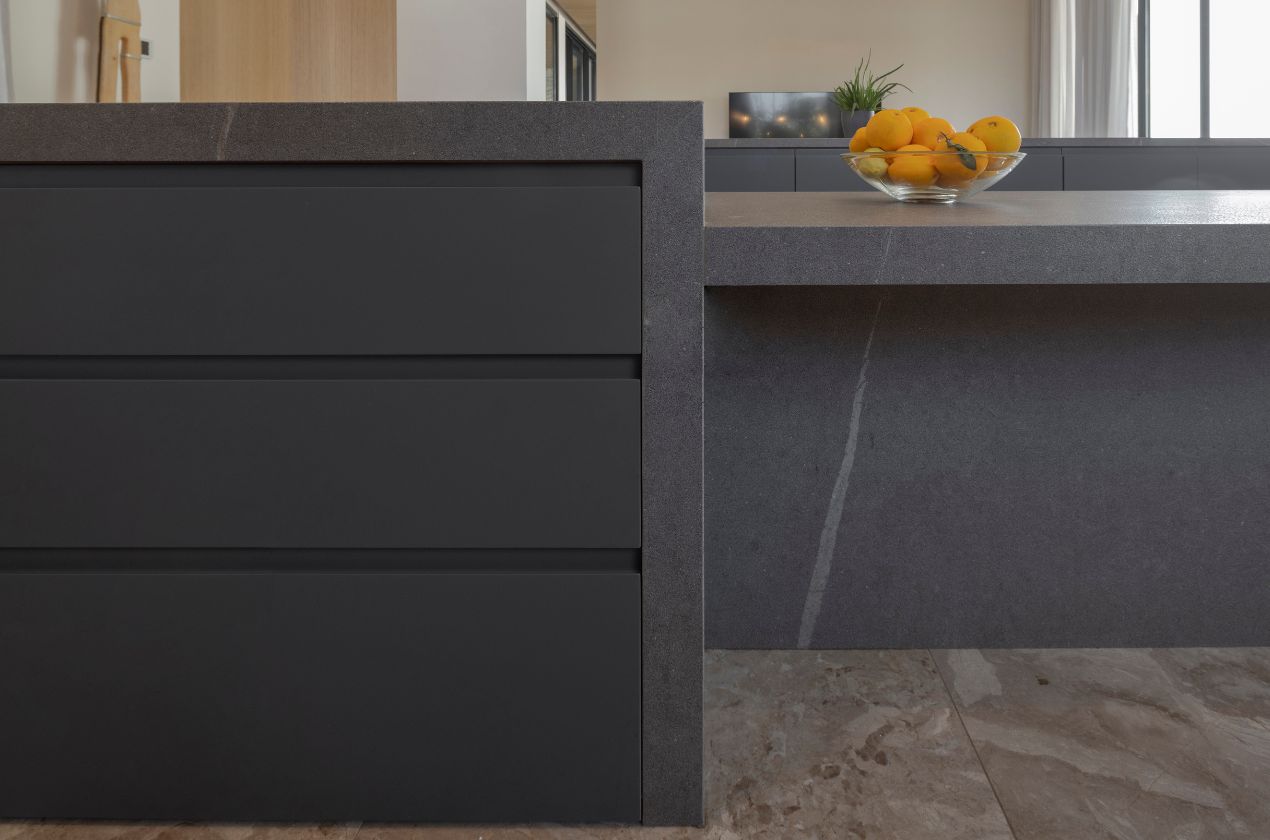 Senda MDi Kitchen Worktop Close-up of a dark gray Senda MDi kitchen Worktop with subtle veining, matte black drawers, and a glass bowl of citrus fruits on a stone-tiled kitchen floor.